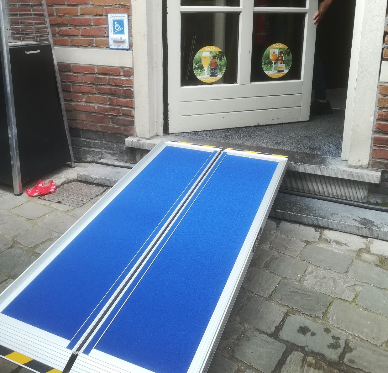 blauwe plooibare antislip oprijplaat die ligt op een 2 trapsdrempel bij een open deur van een winkel  